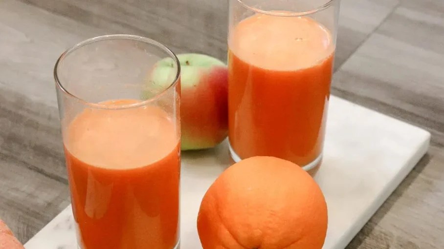 two glasses of orange-colored juice surrounded by an orange and a green apple with red patches